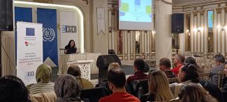 Bir konferans salonu sahnesi  Önde oturan izleyici kitlesi, çeşitli yaşlarda ve cinsiyetlerde insanlar, bazıları başörtülü Sahnenin ortasında masa arkasında oturan, dizüstü bilgisayar kullanan kadın konuşmacı Sol tarafta dikey bir açılır pankart: üstünde Avrupa Birliği bayrağı. Arkada mavi ve beyaz kurul afişleri ve sağda projeksiyon ekranında sunum slaydı Sahnenin önünde büyük siyah hoparlörler ve beyaz süslü bir kürsü bulunuyor.