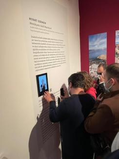 Fotoğrafların bulunduğu kırmızı bir duvarın yanında üzerinde latin alfabesi ile yazılmış yazılar, Braille alfabesi ile yazılmış daha kısa bir metin, bir karekod ve bir işaret dili tercümanının gözüktüğü bir ekran yer alan bilgi panosu var. Panonun önünde bir kadın, iki erkek dokunarak ve yakından bakarak panoda ne olduğunu keşfediyor.