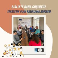 Görselde, bir masa etrafında toplanmış bir grup insan görülüyor. Masada dizüstü bilgisayarlar ve not defterleri var. Arka planda "Yerel STÖ'ler Kurumsal Destek Programı" yazılı bir afiş asılı. Afişte Avrupa Birliği ve STGM logoları yer alıyor. Görselin üst kısmında "Birlikte Daha Güçlüyüz" ve "Stratejik Plan Hazırlama Atölyesi" yazıları bulunuyor. Alt kısımda ise EGED logosu ve bazı grafik tasarımlar var. Görselin genel havası, bir toplantı veya atölye çalışması yapıldığını gösteriyor.