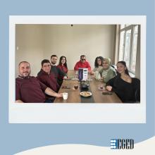 Bir toplantı masası etrafında oturan bir grup resmi  Ön planda soldan sağa: •Koyu bordo uzun kollu tişört giymiş, sağında bir beyaz fincan tutan sakallı erkek (gülümseyen) •Bordo kazaklı, gözlüklü genç erkek •Koyu sweatshirt giymiş sakallı genç erkek •Uzun koyu saçlı, kırmızı üstlü genç kadın  Masanın ortasında: •Gözlük takan kırmızı üstlü erkek (elleri masada) •Masada bir dikey duran beyaz-lacivert masa tabelası veya reklam kartı görünüyor.