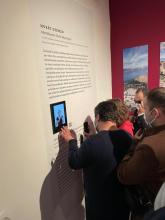 Fotoğrafların bulunduğu kırmızı bir duvarın yanında üzerinde latin alfabesi ile yazılmış yazılar, Braille alfabesi ile yazılmış daha kısa bir metin, bir karekod ve bir işaret dili tercümanının gözüktüğü bir ekran yer alan bilgi panosu var. Panonun önünde bir kadın, iki erkek dokunarak ve yakından bakarak panoda ne olduğunu keşfediyor.